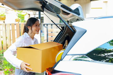 Beautiful asian businesswoman driving car parked in front of her house in a good mood holding cardboard box walking into new residence moving in on a sunny moving day looking happily at the camera.