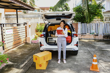 Beautiful asian businesswoman with successful business, preparing to move moving luggage in trunk holding clipboard notes list of things with bright smile good mood : Relocation begins with new home.