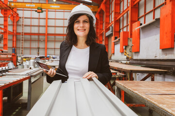 Female manager engineer wearing safety helmet inspects the production quality of white coated rolled metal roofing sheets, in a standardized and safe warehouse production facility.