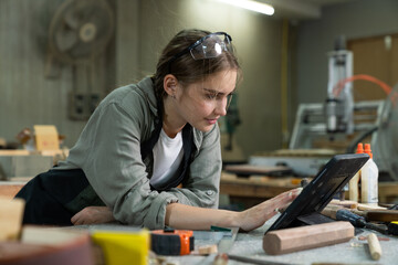 A young woman is training to be a carpenter in the workshop. smartphone and working with a laptop...