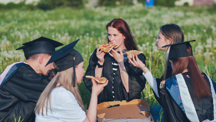 Graduates in black suits eating pizza in a city meadow.