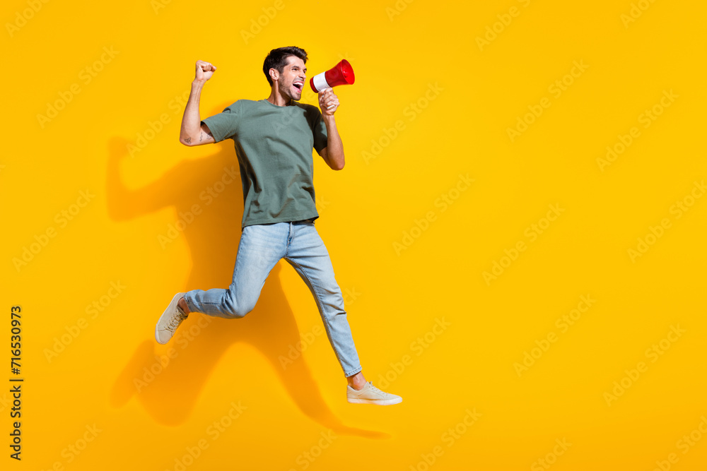 Poster full length photo of cool lucky man dressed khaki t-shirt rising fist shouting speaker jumping high 
