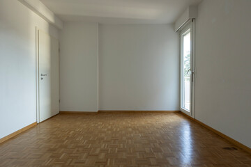 Front view of an empty room with parquet flooring and white walls. On the left is a window letting in a lot of natural light, on the right is a closed door.