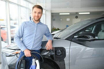 Concept of buying electric vehicle. Handsome business man stands near electric car at dealership