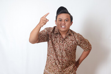 Portrait of unpleasant Asian man wearing batik shirt and songkok showing cynical unhappy angry facial expression pointing forward, giving warn. Isolated image on gray background - obrazy, fototapety, plakaty