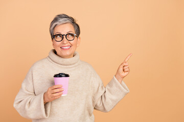 Portrait of charming cheerful lady hold coffee cup look direct finger empty space isolated on beige color background