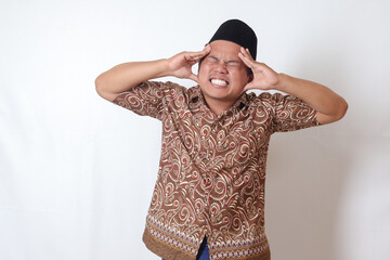 Portrait of suffering Asian man wearing batik shirt and songkok having headache while touching his forehead area. Isolated image on gray background