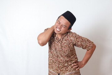 Portrait of suffering Asian man wearing batik shirt and songkok having neckache while touching his back. Isolated image on gray background