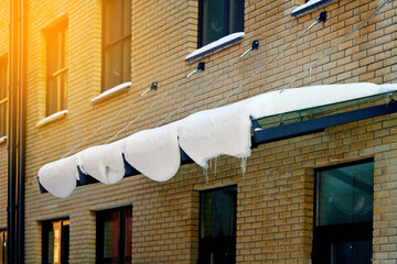 Door canopy with melting snow and ice in sunny day, danger for pedestrians. Glass panel mounted with tension rod and wall brackets on building facade. Snow melts on the awning in front of the door