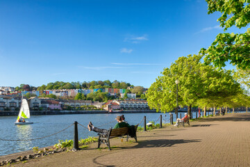 Bristol, UK - A beautiful spring evening at Bristol Docks, young couple sitting on a bench, colourful houses, fresh green trees, blue sky, sail boat on the water.