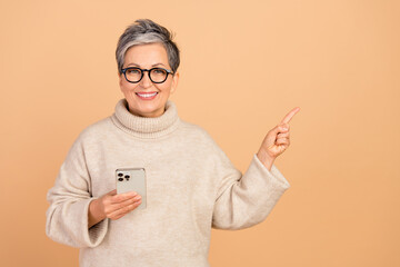 Portrait of grandmother in white turtleneck directing forefinger empty space apple iphone new model isolated on beige color background