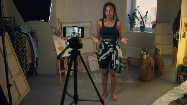 Full length shot of backstage of young Black woman filming herself on phone using tripod and introducing on camera then showing dance moves at home
