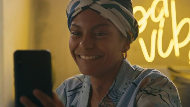 Handheld closeup of pretty young Black woman in durag smiling while speaking during video call using smartphone in modern room with neon sign in evening