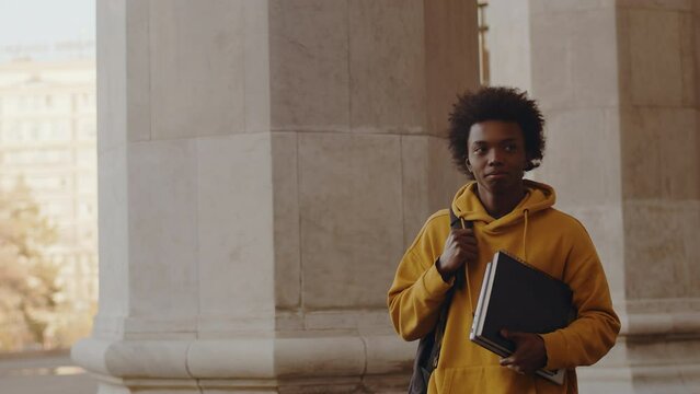 Young black programmer walking alongside the building outdoors after a successful working day with a smiling satisfied face holding a laptop and copybook