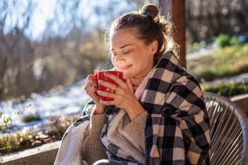 Young woman enjoying a sunny early spring morning on the terrace, wrapping herself in a blanket and...