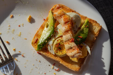 A healthy breakfast of toast topped with boiled egg, avocado, and grilled chicken breast, served on a white plate bathed in sunlight from a window.