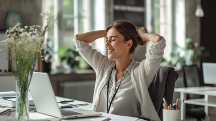 Woman with her hands behind her head is relaxing at her office desk , a smile on her face, and a laptop in front of her. - obrazy, fototapety, plakaty