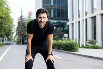 Portrait of tired young man standing bent over on street road after jogging and breathing hard, resting after marathon and training - obrazy, fototapety, plakaty