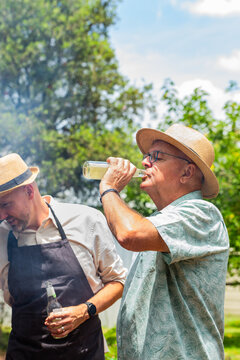Man in his sixties drinking cider from bottle while standing by barbeque with his adult son
