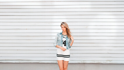 beautiful young fashionable girl in a trendy white skirt with a denim jacket stands near a vintage...