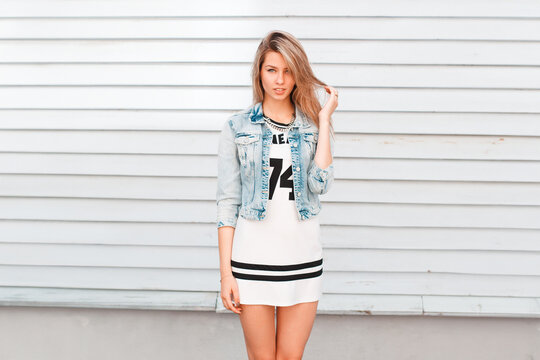 Beautiful young woman in fashion street denim clothes with a dress and a jeans jacket stands near a vintage wooden wall