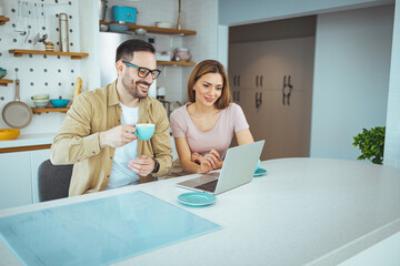 Pleasant family couple sitting at big wooden table in modern kitchen, looking at laptop screen....