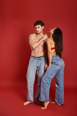 muscular and shirtless man in jeans standing near gorgeous brunette woman in bra on red backdrop
