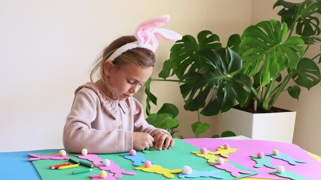 Child girl in rabbit ears creating Easter paper colored garland Easter bunny. Cute children's crafts for Easter party at home. Holiday Art Activity Kid. Fine motor skills, creativity and hobby DIY
