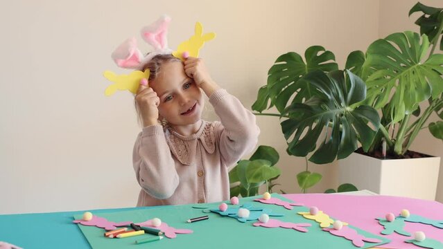Happy child girl in bunny ears make and play Easter paper colored garland Easter bunny. Children crafts for Easter party at home. Holiday Art Activity Kid. Fine motor skills, creativity and hobby DIY