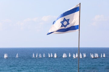 Israeli flag flutters in the wind, the open sea, and sailboats in the background.
