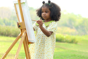Cute smiling African girl with black curly hair painting on canvas at green garden outdoor. Kid...