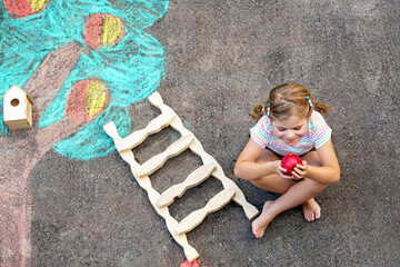 Fototapeta premium Cute little girl painting with colorful chalks apples harvest from apple tree on asphalt. Cute preschool child with having fun with chalk picture. Creative leisure for children, drawing and painting.