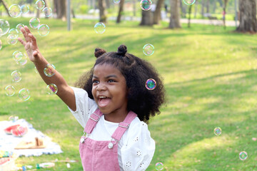 Happy smiling African girl with black curly hair blowing and playing with soap bubbles at green...
