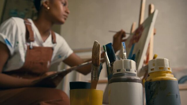 Closeup of paints and multiple paintbrushes in cups with female African American artist creating artwork in background in studio