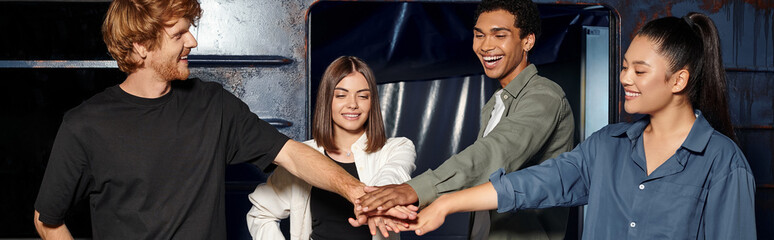 young multicultural group of friends stacking hands while having quest in escape room, banner