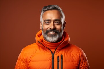 Obraz premium Portrait of a jovial indian man in his 50s donning a durable down jacket against a soft orange background. AI Generation