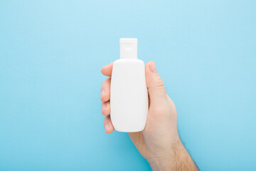 Young adult man hand holding white plastic cream bottle on light blue table background. Pastel...