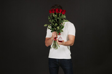 Handsome young man is posing on grey background with gift box and red roses in hands, looking at camera. Happy Valentine`s day.