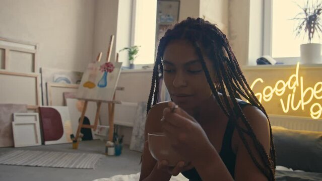 Tilt closeup of young Black lady with box braids using laptop sitting on floor mattress and eating meal near dog in modern grunge loft apartment