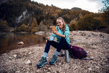 Happy female hiker drinking coffee and using smartphone by the lake