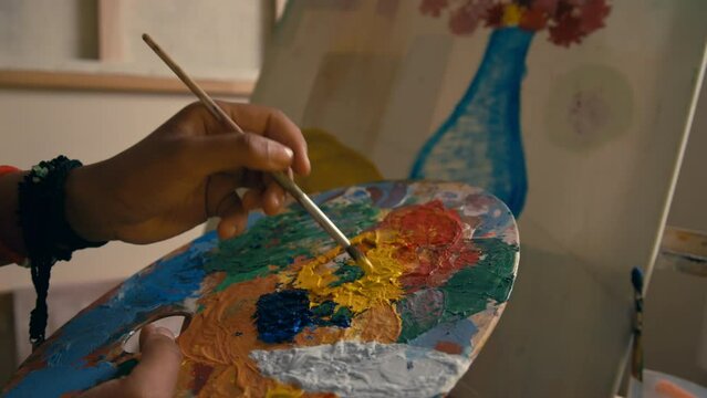 Closeup of unrecognizable left-handed artist holding palette with multiple bright colors of paint while creating new artwork in studio
