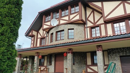 Modern private three-story country house in English, European style with manicured green grass, stone wall, thuja fence and red tile roof. Pseudo-historic style of architecture.