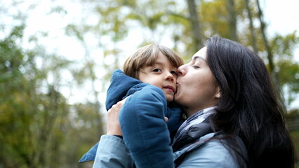 Fototapeta premium Mother lifting on in arms and kissing in cheek standing outside during autumn fall season, authentic lifestyle family scene of mom and child loving relationship