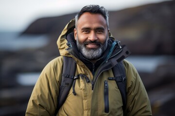 Fototapeta premium Portrait of a grinning indian man in his 40s donning a durable down jacket against a rocky shoreline background. AI Generation