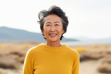 Portrait of a grinning asian woman in her 70s showing off a thermal merino wool top against a serene dune landscape background. AI Generation