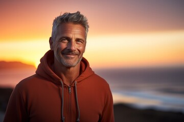 Portrait of a smiling man in his 50s wearing a zip-up fleece hoodie against a vibrant beach sunset...