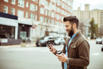 Obraz na płótnie Canvas Man with beard using smartphone and holding coffee cup on city street