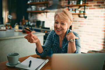 Smiling senior woman holding credit card looking at laptop in home kitchen