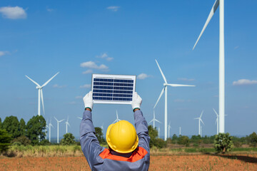 A man engineer in hard hat is holding solar panels in wind farm, clean renewable energy concept
