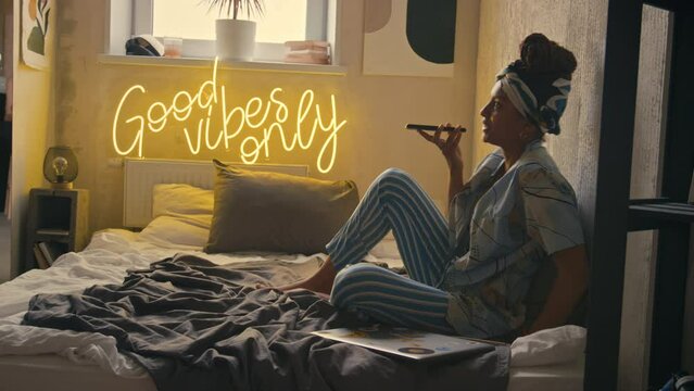 Full side shot of young stylish Black woman in headband recording audio message with smartphone sitting in bed in modern grunge room with neon caption Good Vibes Only on wall